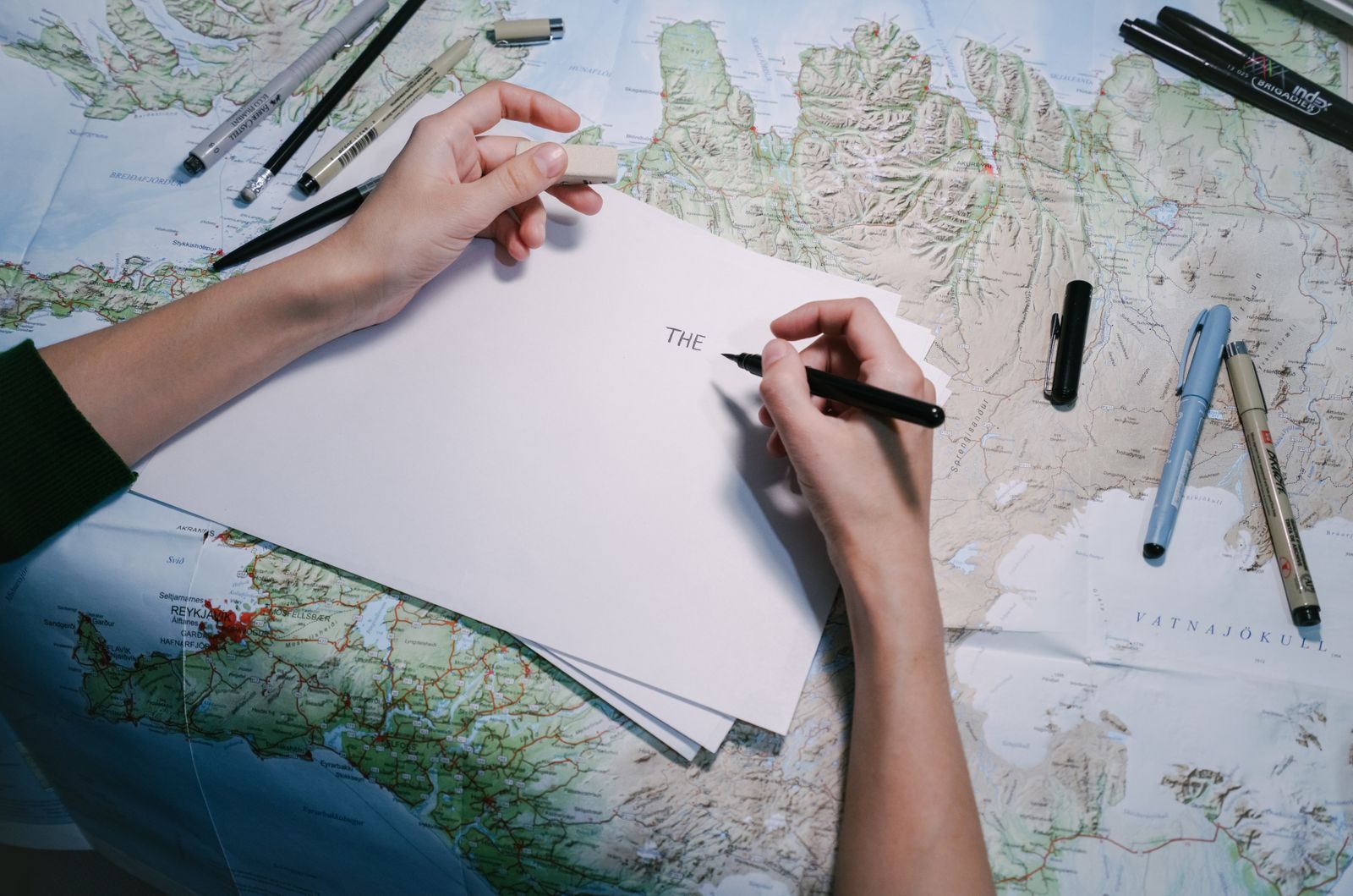 Hands writing a travel question on a sheet of paper laid over a map, pens alongside