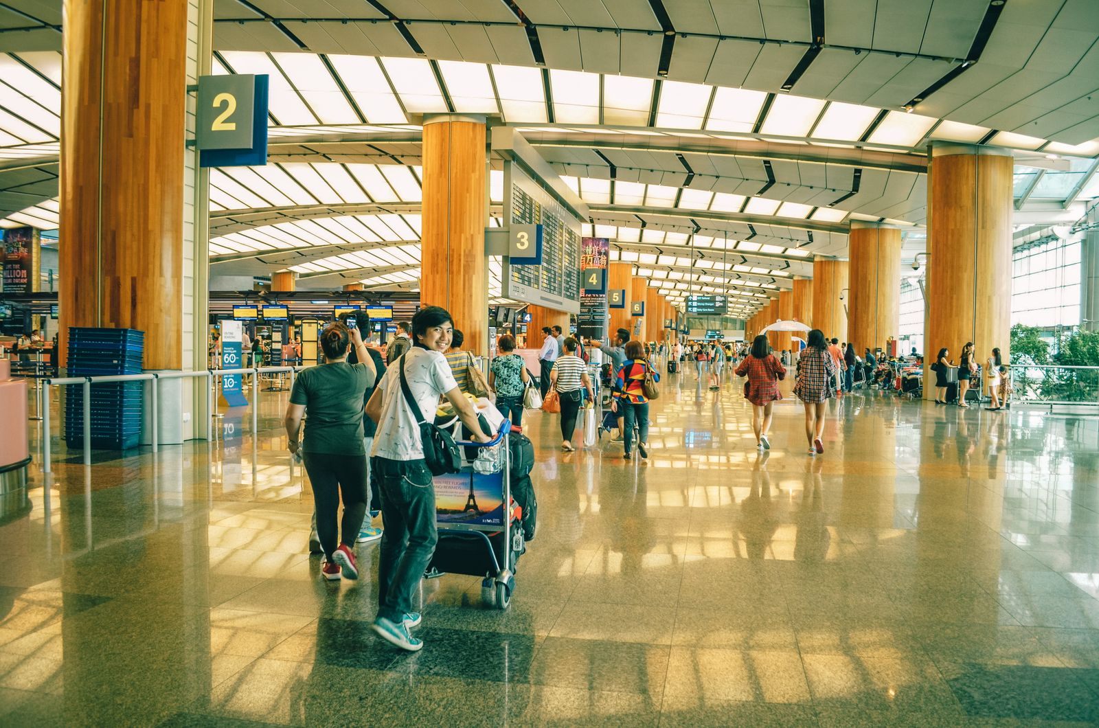 Travelers with a luggage cart crossing a bright, busy airport terminal hall