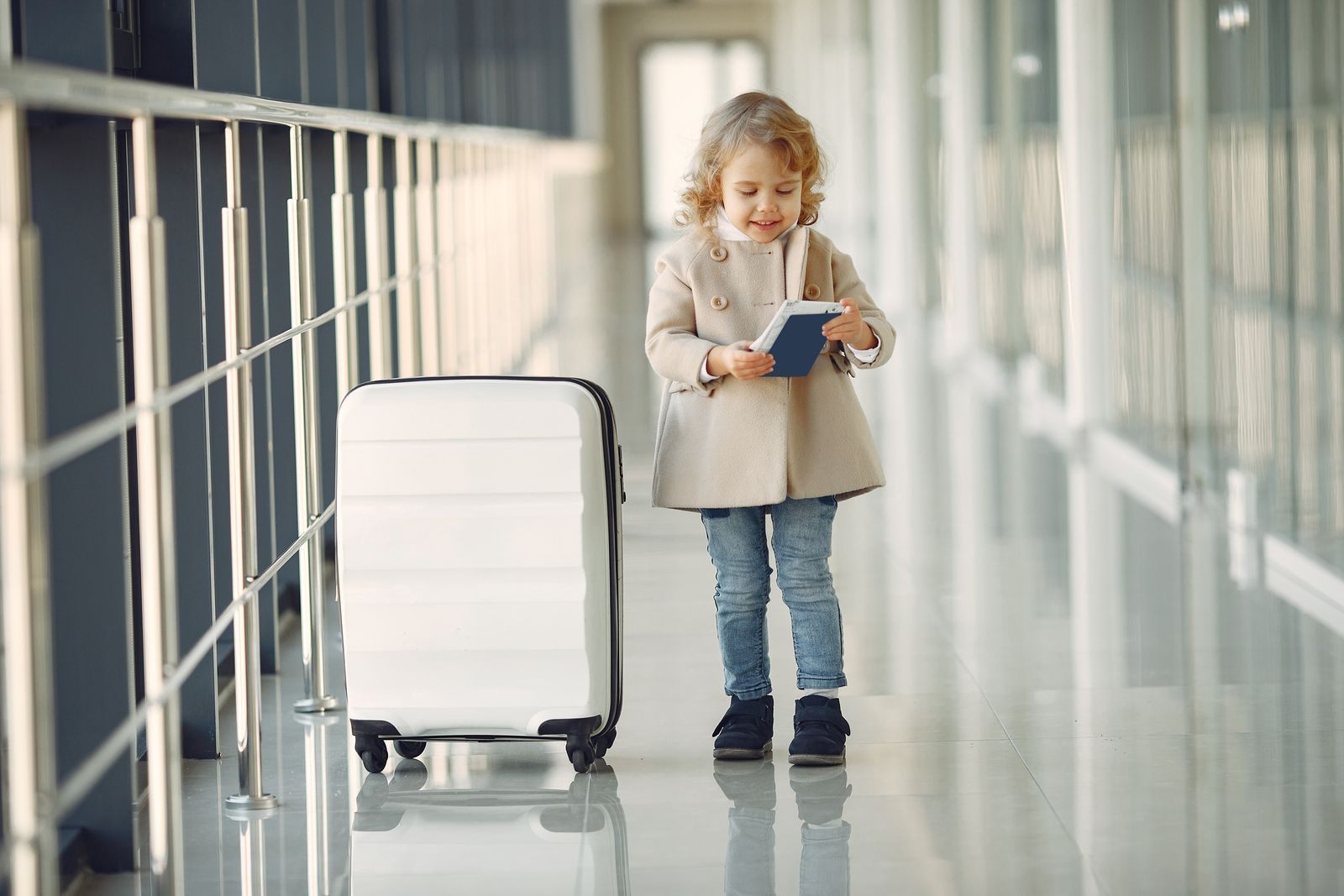 Little girl holding a passport next to a white suitcase in a glass airport corridor