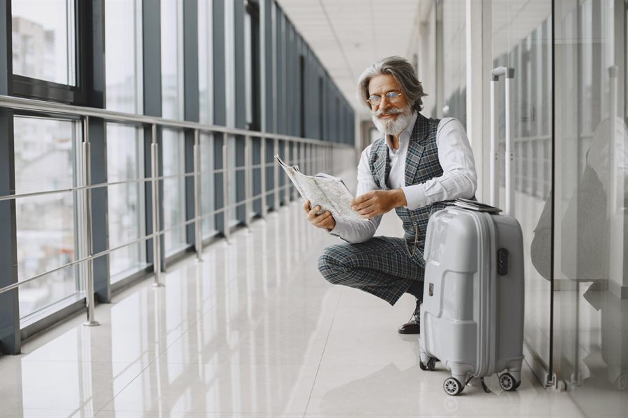 Older traveler crouching beside a hard-shell suitcase in an airport walkway, reading a printed map